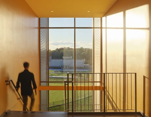 A person descends a staircase framed by large windows, revealing a vibrant view of a sports field bathed in golden sunlight.