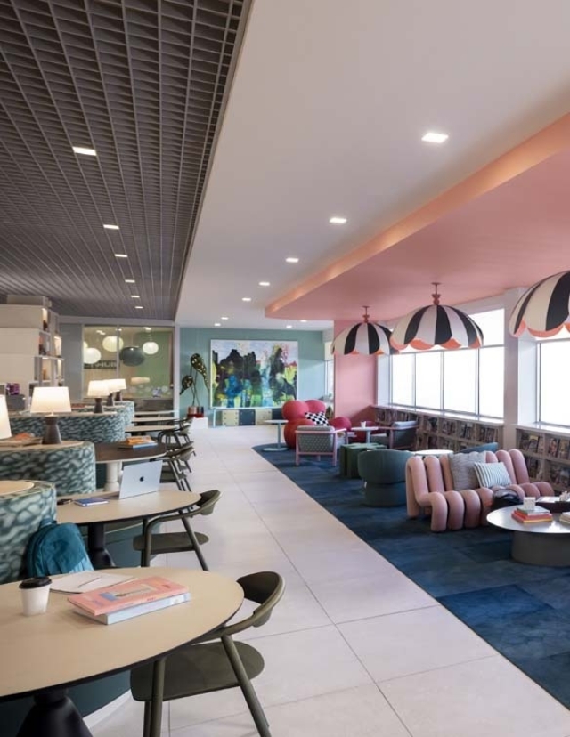 A modern, vibrant lounge featuring colorful furniture, a large artwork, and cozy seating arrangements, illuminated by natural light.