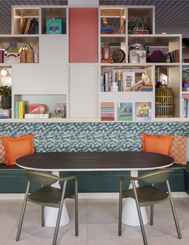 A modern dining area features a sleek oval table surrounded by two minimalist chairs, with a vibrant bookshelf and cushioned seating.