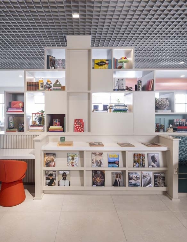 A modern bookshelf features colorful decor and neatly arranged magazines, alongside a vibrant red stool in a stylish, airy space.