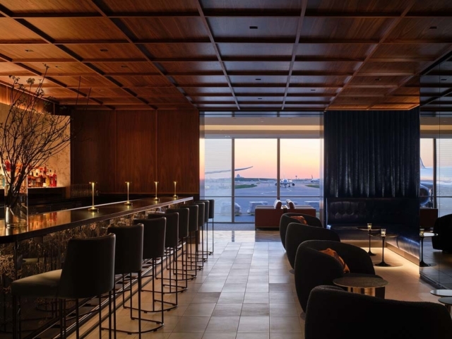 A sleek airport lounge features a long, stylish bar with stools, cozy seating areas, and large windows showcasing a sunset view.