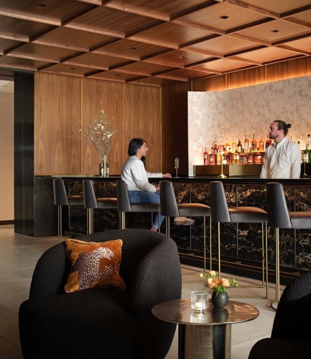 A stylish indoor bar features a bartender serving a seated customer. The décor includes sleek wood paneling and elegant lighting.