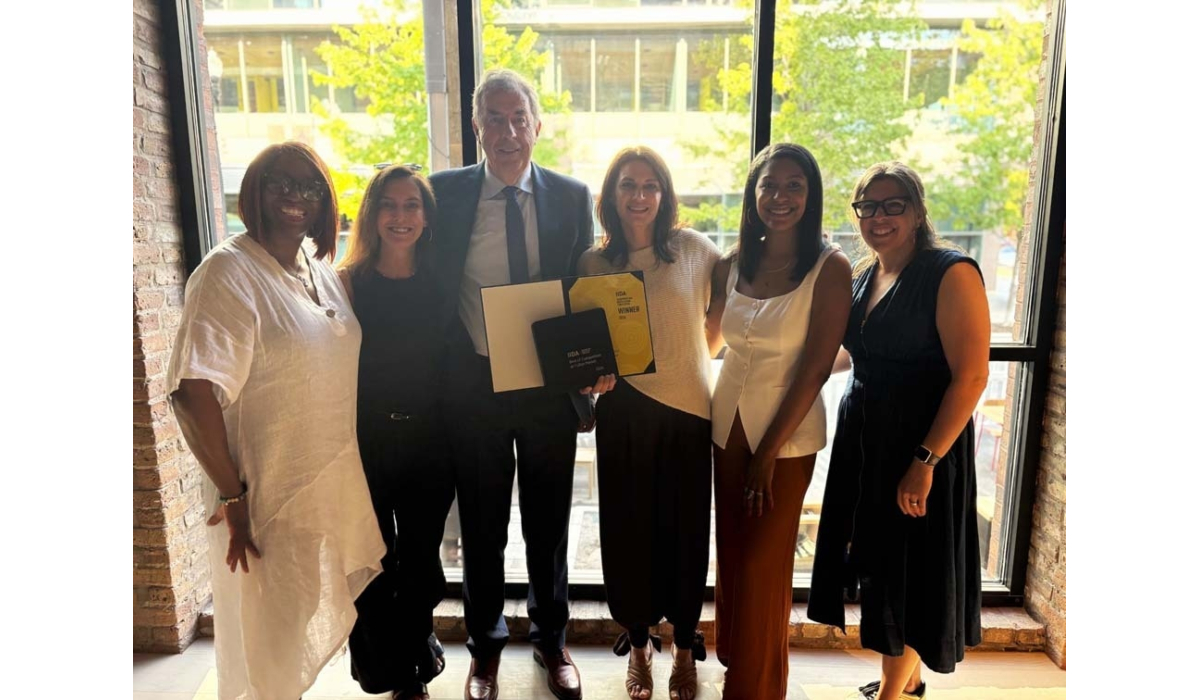 Six smiling colleagues stand in front of a tall window in daylight; center person holds an award folder while others pose together.