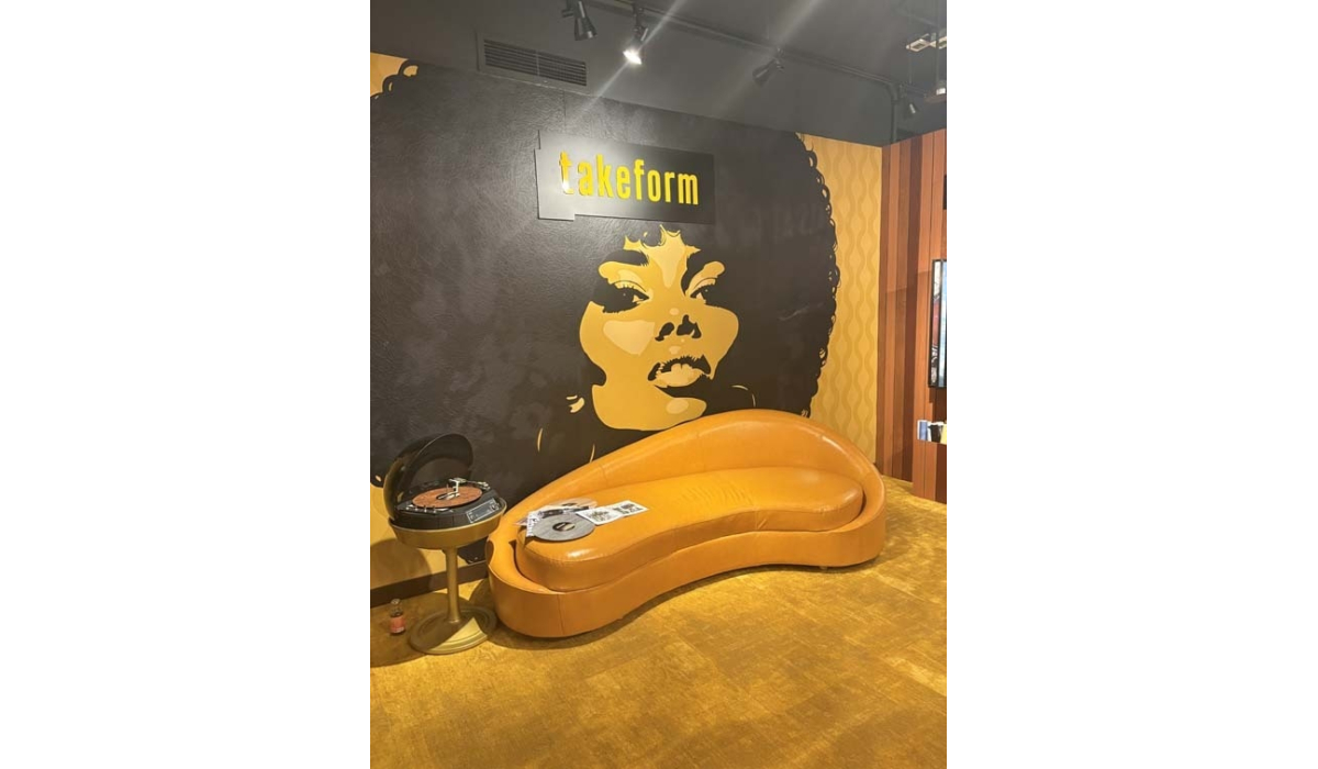 Curvy orange sofa beside a spinning record player and magazine below a black-and-gold mural of an Afro woman's face with a 'takeform' sign.