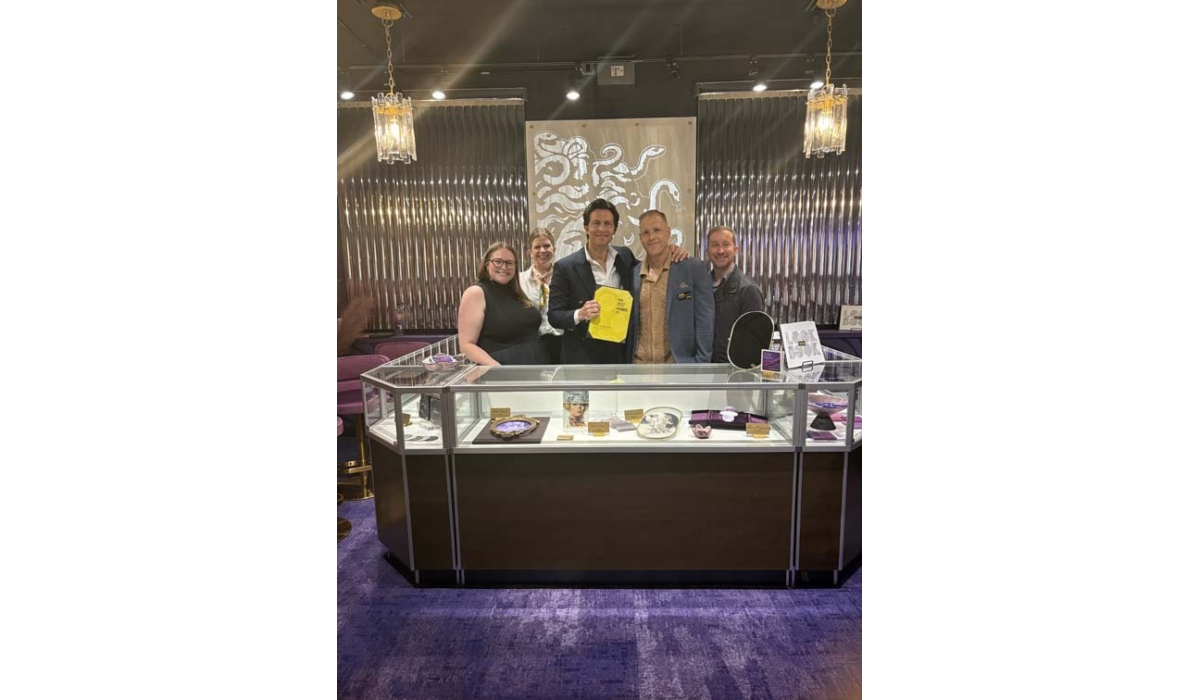 Five smiling staff stand behind a glass case of jewelry, purple carpet and metallic octopus wall art; center man holds a yellow bag.