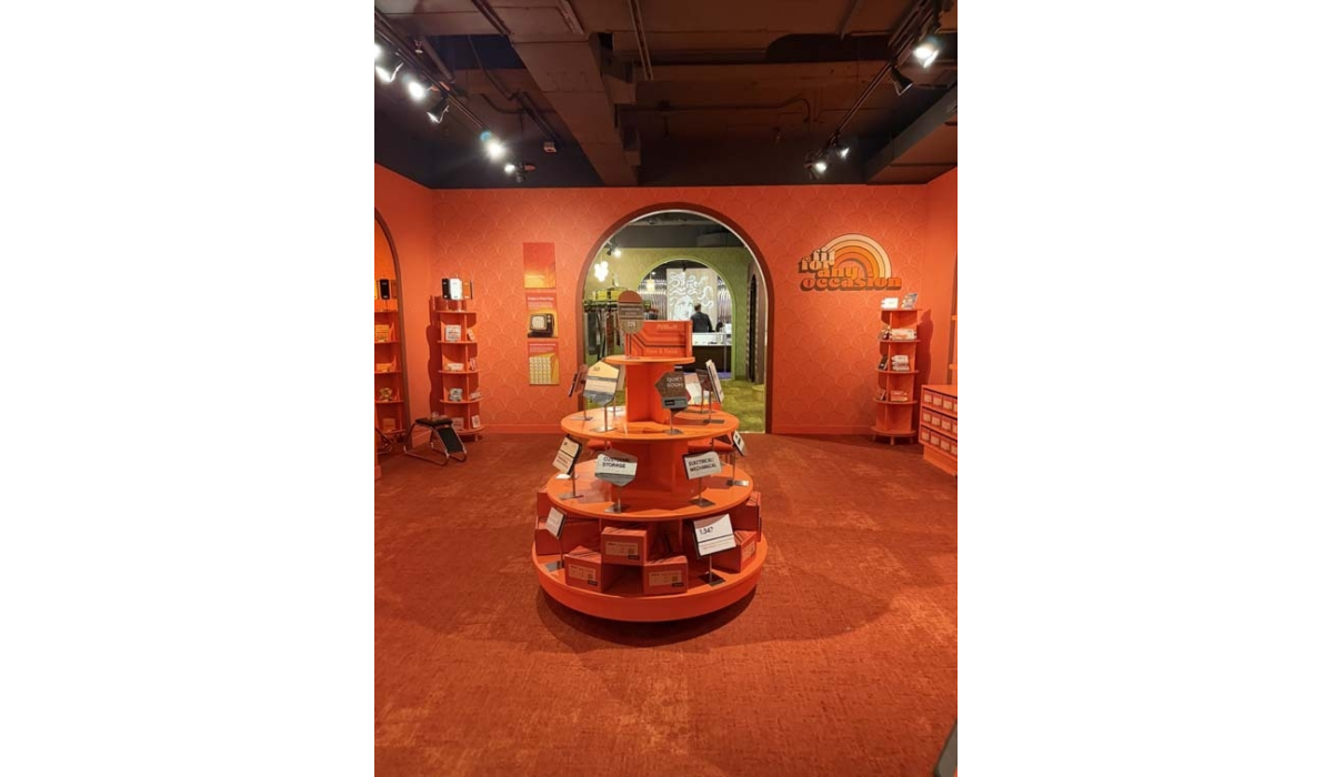 Retro orange-red boutique interior with a circular tiered display of boxed goods centered on carpet, arched doorway revealing green room.