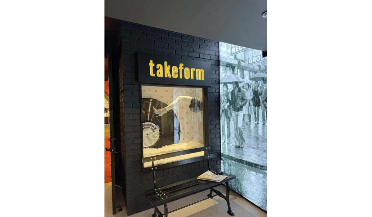 Black brick storefront labeled 'takeform' with a mannequin leg and vinyl record display behind glass, bench and vintage newspaper nearby.