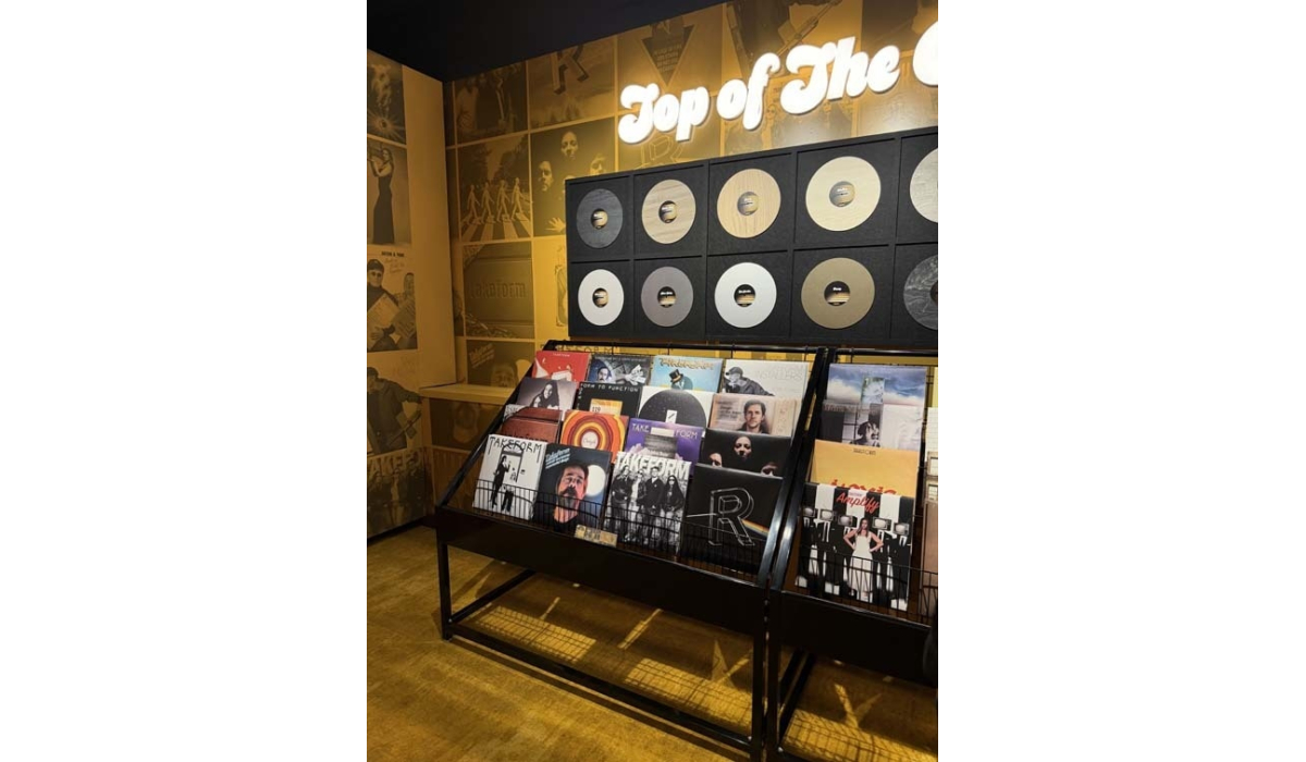 Vinyl display in warm yellow lighting: black metal racks holding albums beneath framed gold records and neon 'Top of The' sign.