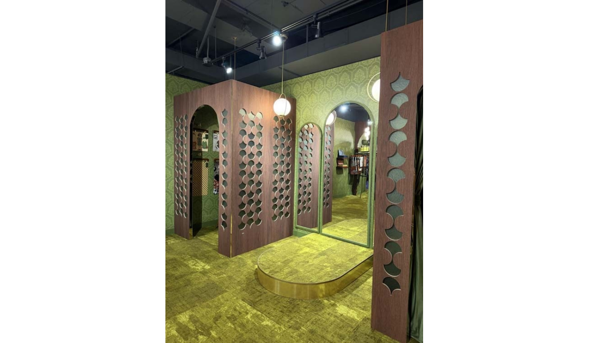 Retro fitting room with green wallpaper, gold carpet, arched mirror, wooden panels with circular cutouts and globe lights and racks.