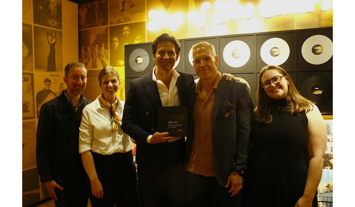 Five smiling adults stand in a warmly lit room with retro records on the wall, the center man holding a small award plaque flanked by peers.