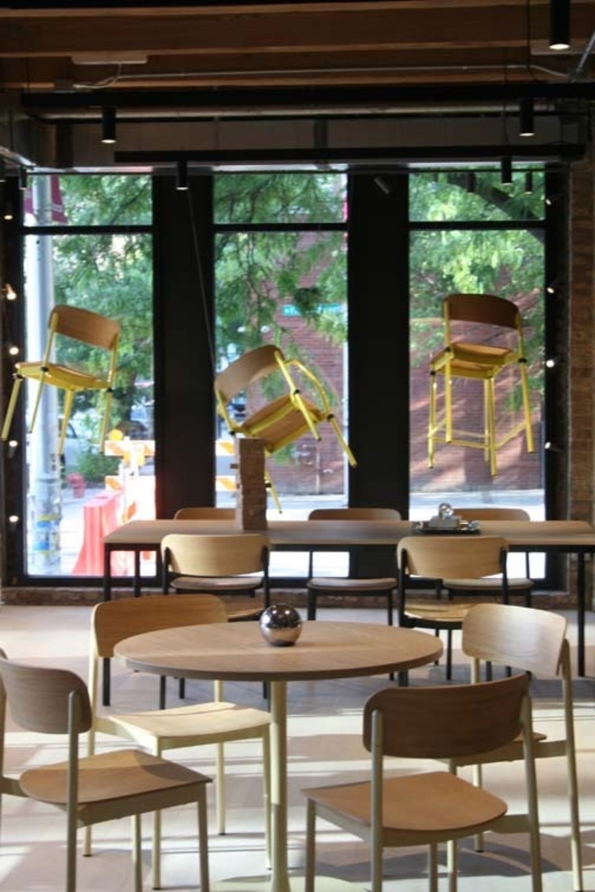 Modern café interior with round wooden tables and chairs, 3 yellow bar stools suspended in front of large windows overlooking leafy street.