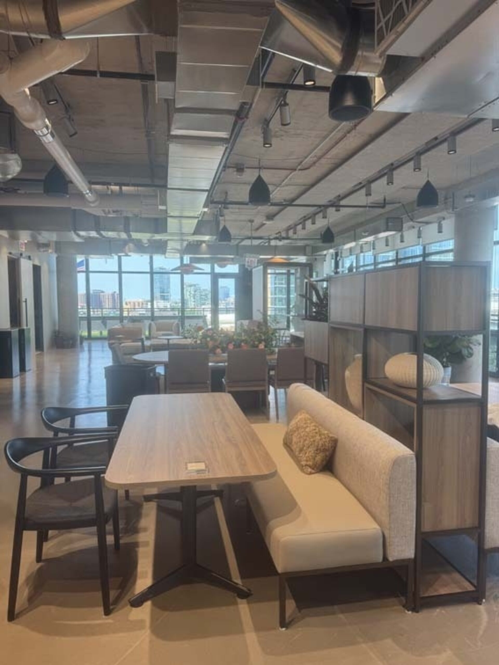 Modern coworking lounge with wooden table, bench seating, chairs, open shelving, pendant lights and large windows showing city skyline.