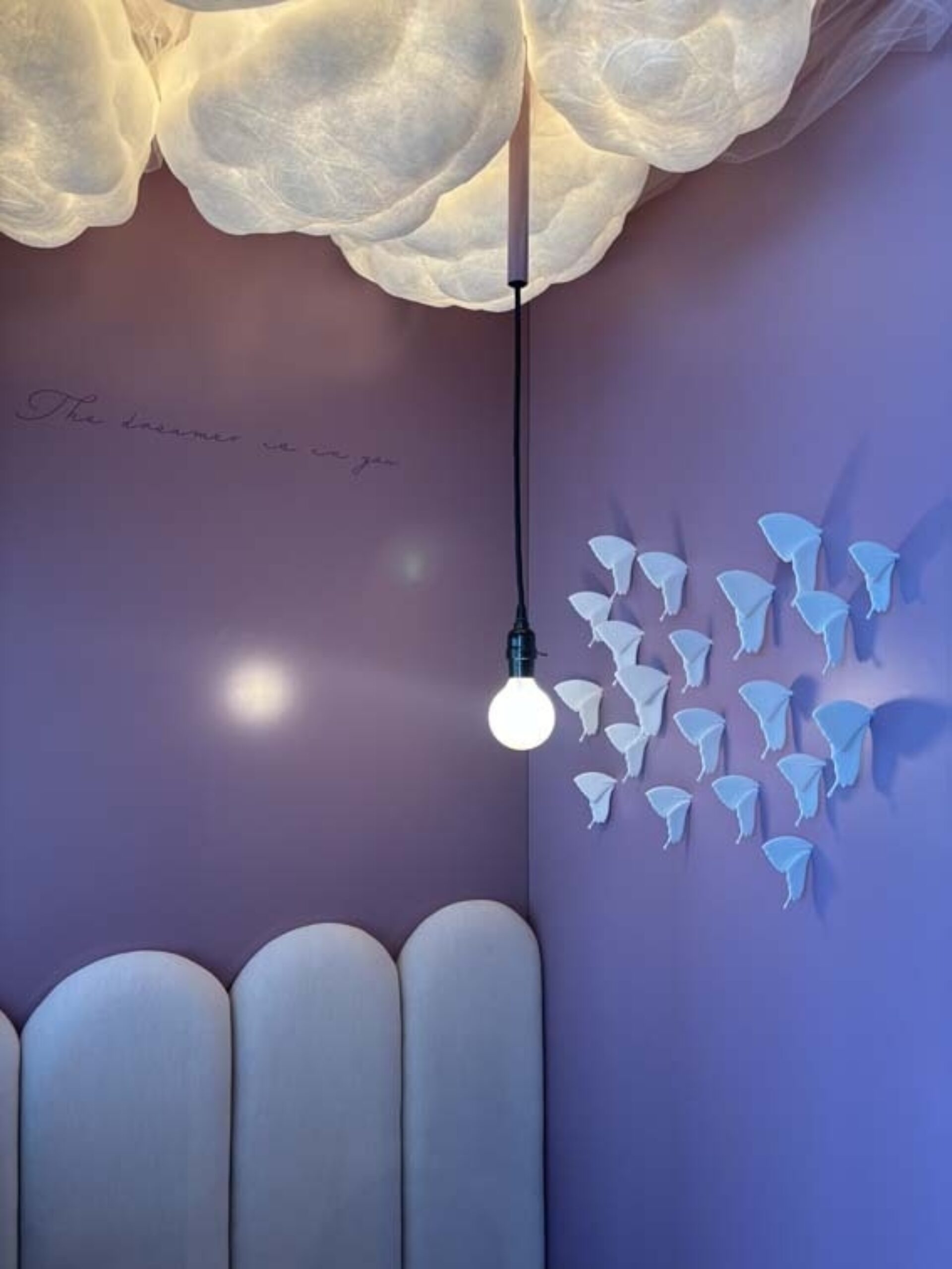 Lavender corner with cloudlike ceiling lights, a single hanging bulb above a scalloped cream headboard and white 3D butterfly wall art.