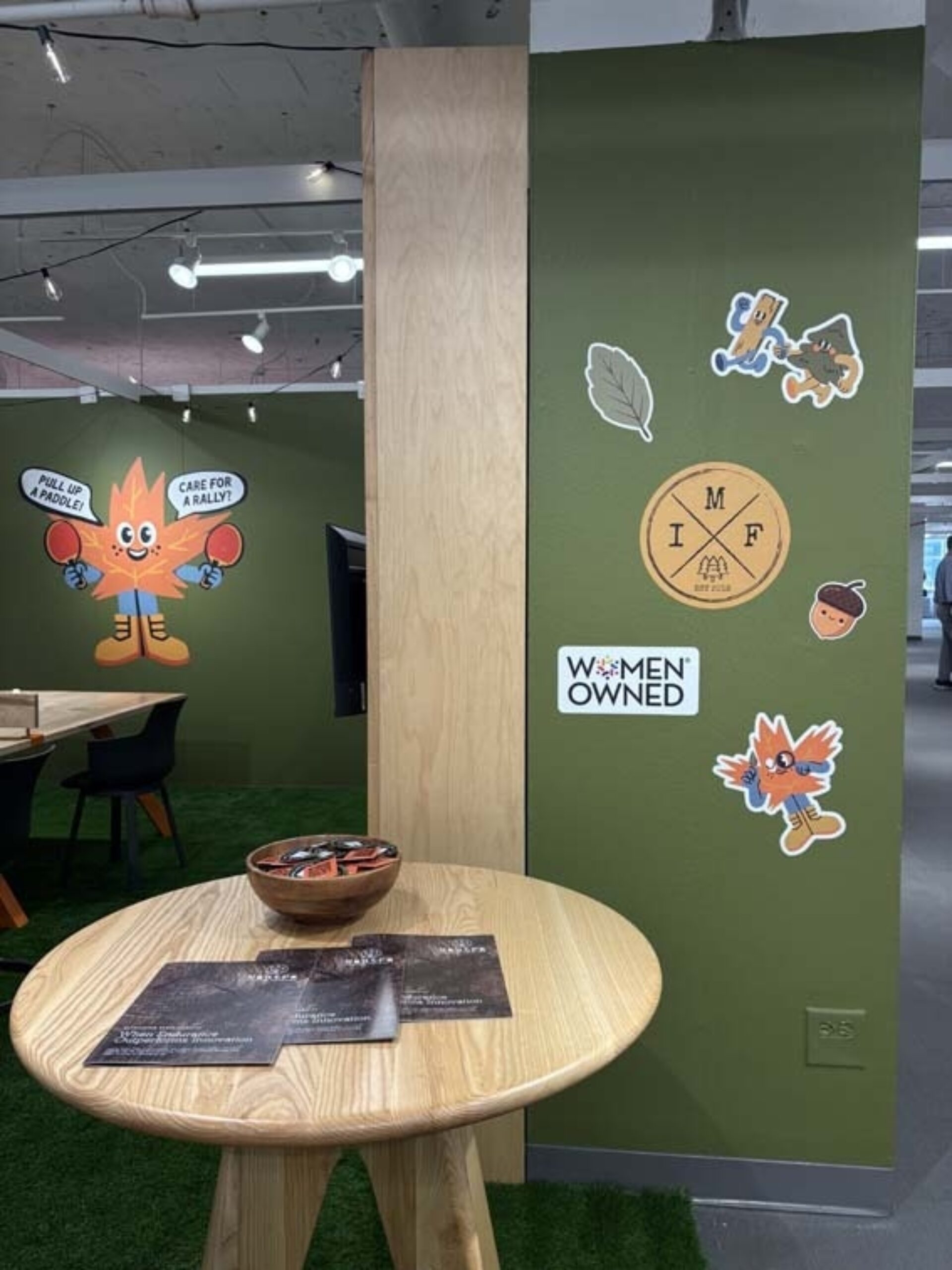 Office corner with olive-green walls and playful maple-leaf mascots, a Women Owned sticker, and a round wood table with snacks and flyers.