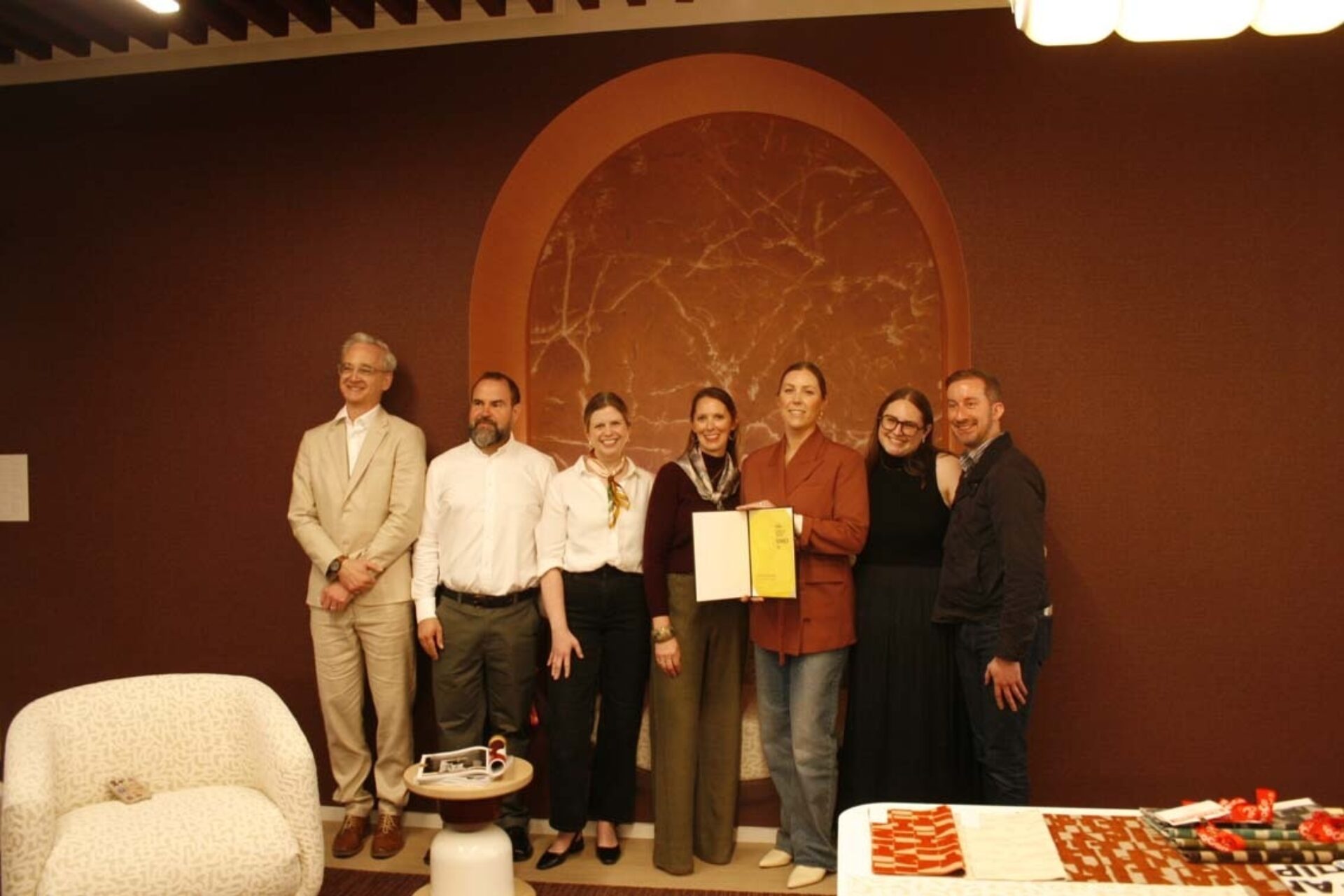 Seven people stand in a warm, brown room beneath an arched marble niche, smiling as a woman near center holds a yellow certificate.