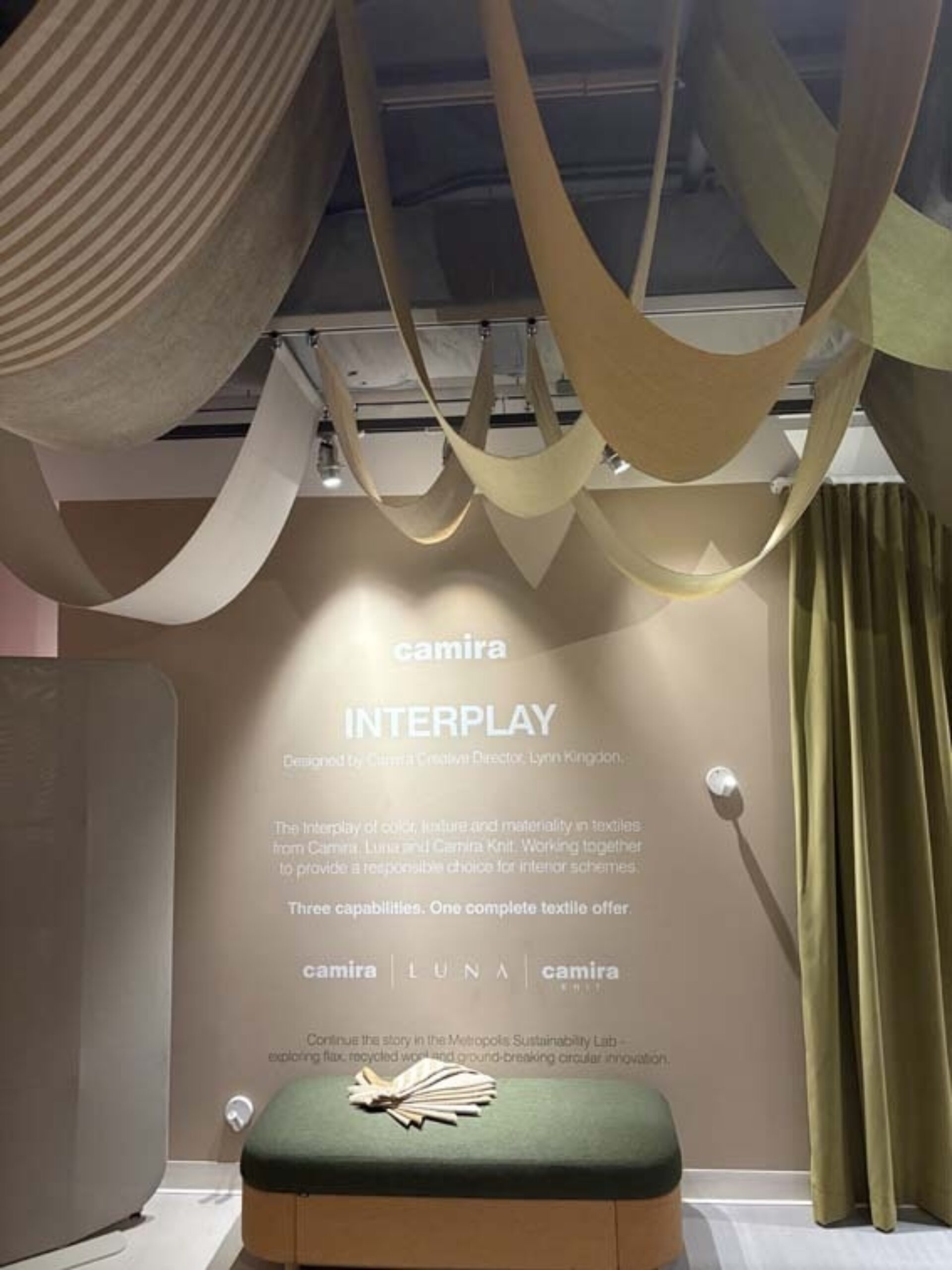 Hanging undulating textile swags in beige, cream and olive above a muted pink wall with 'camira INTERPLAY' text, green bench and curtain.