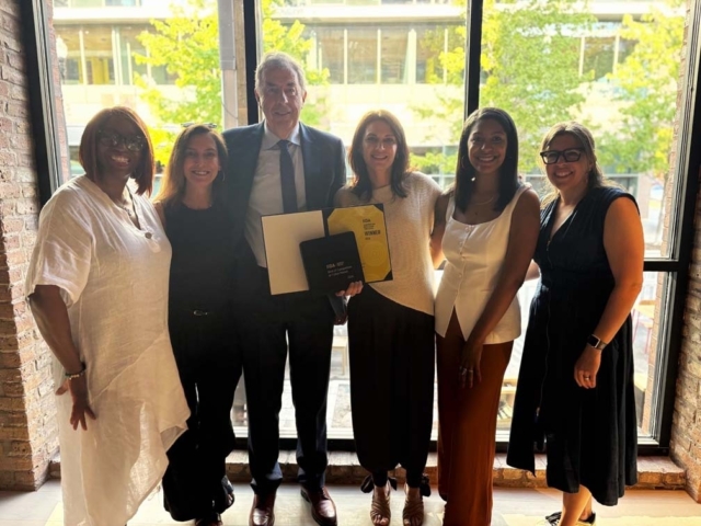 Six smiling colleagues stand in front of a tall window in daylight; center person holds an award folder while others pose together.
