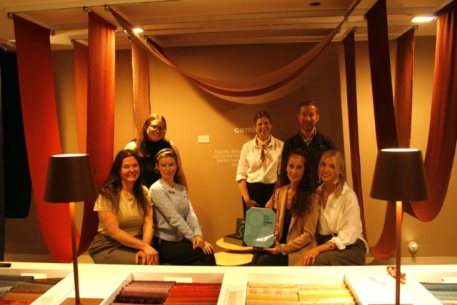 Seven designers pose in a warm textile showroom with rust-red hanging fabric samples, two table lamps and a small award held at center.