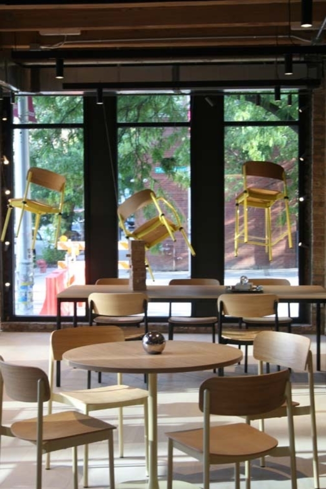 Modern café interior with round wooden tables and chairs, 3 yellow bar stools suspended in front of large windows overlooking leafy street.