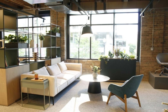 Bright industrial lounge with large windows and exposed brick, white sofa, teal lounge chair, round black coffee table and plants.
