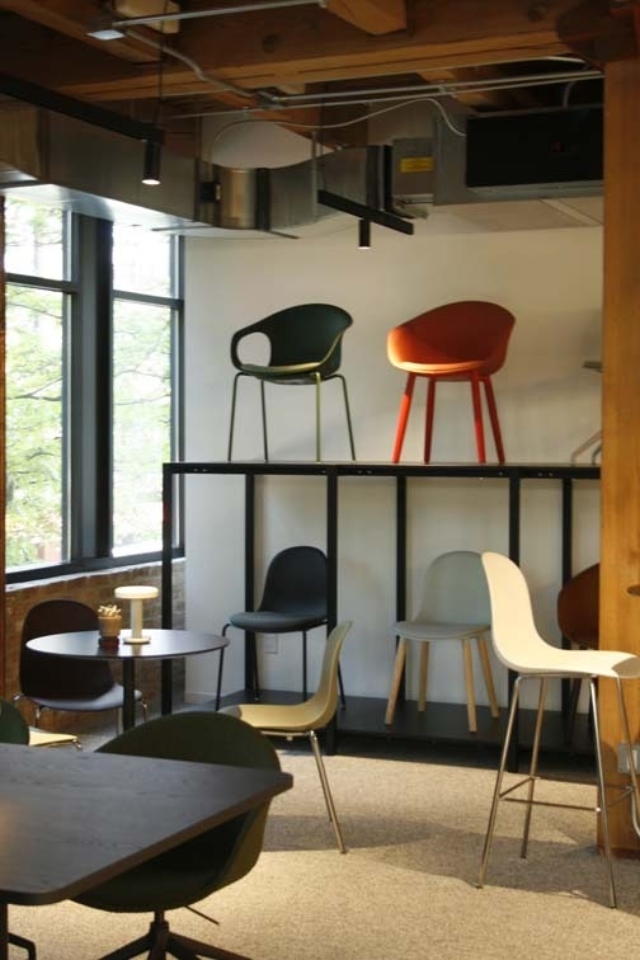 Modern showroom with assorted colorful chairs arranged on black shelving and around tables beneath exposed wooden beams and large windows.