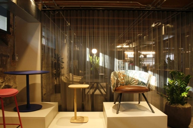 Contemporary showroom corner with a patterned tropical armchair on a pedestal, colorful side tables, sheer curtains and a potted plant.