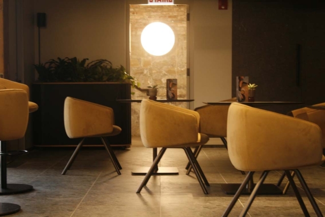 Cozy dim-lit cafe lounge with mustard-yellow upholstered swivel chairs around low black tables, glowing round pendant over a stone doorway.