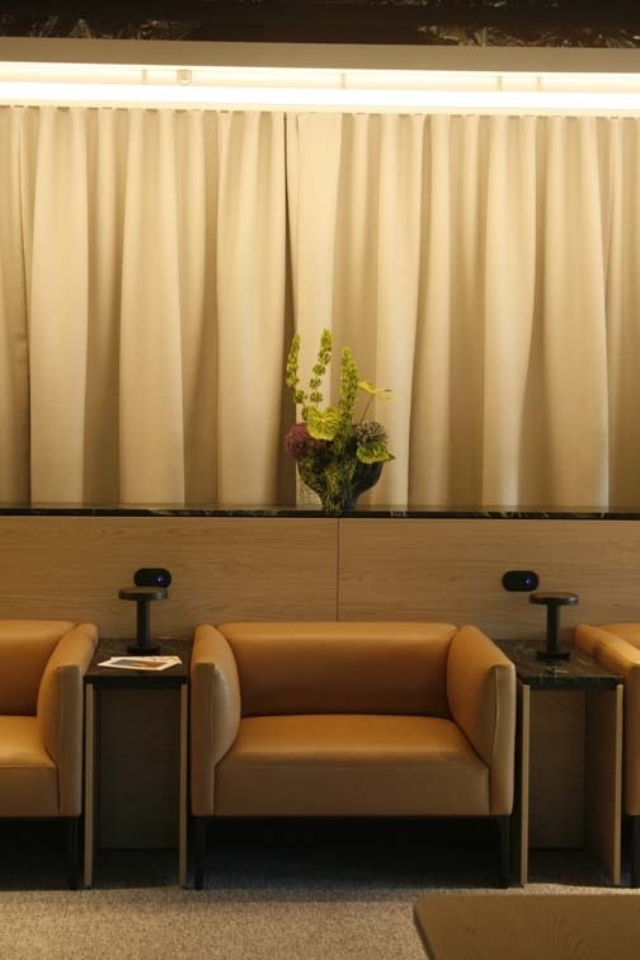 3 tan leather armchairs separated by dark wood side tables with small black lamps, a central shelf holds a vase of green and purple flowers.