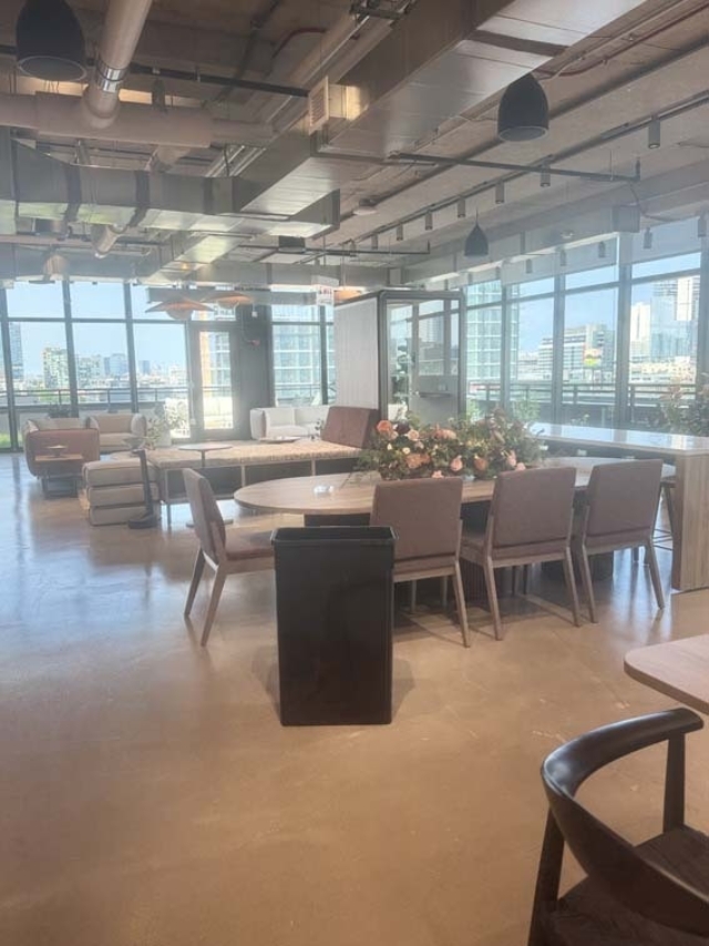 Open office lounge with round wooden table and flower arrangement, brown chairs, sofas near tall windows and exposed ductwork.