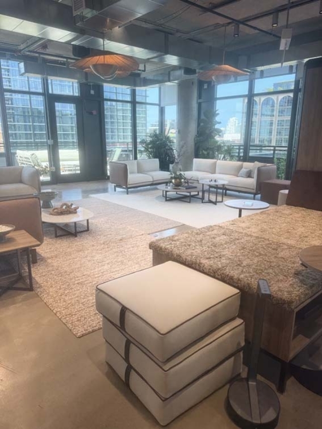 Sunlit lounge with floor-to-ceiling windows, sofas, stacked ottomans, woven pendant lights, layered rugs, round tables and city views.