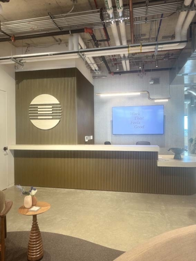 Modern reception with a long ribbed green desk, round slatted logo, wall screen reading Work That Feels Good, and exposed ceiling pipes.