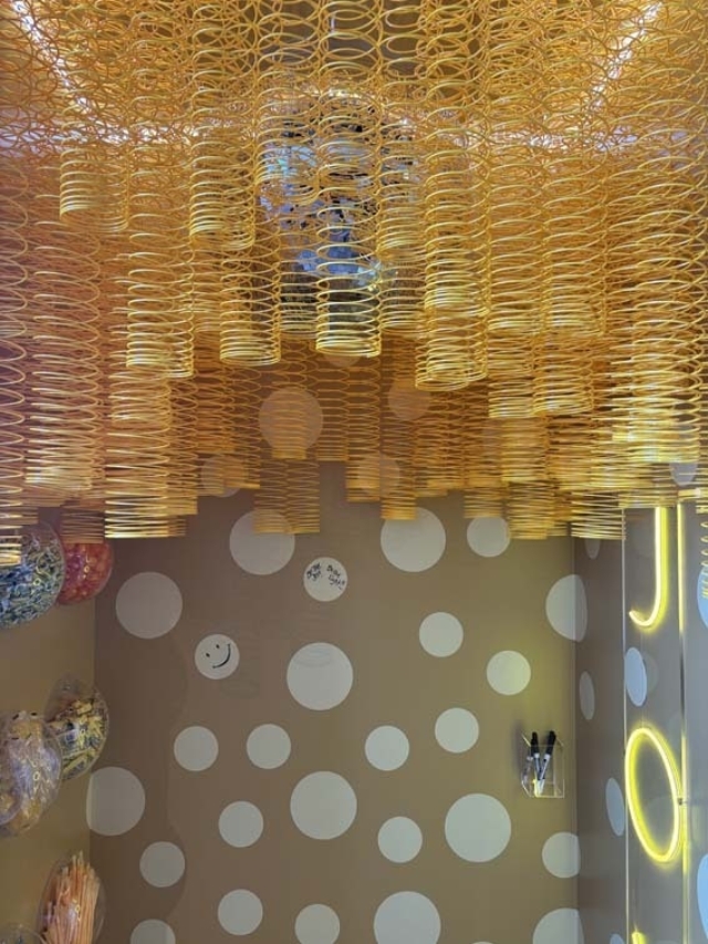 Golden spiral coils hang from the ceiling above a beige wall with large white polka dots, neon accents, balloons and a smiley sticker.