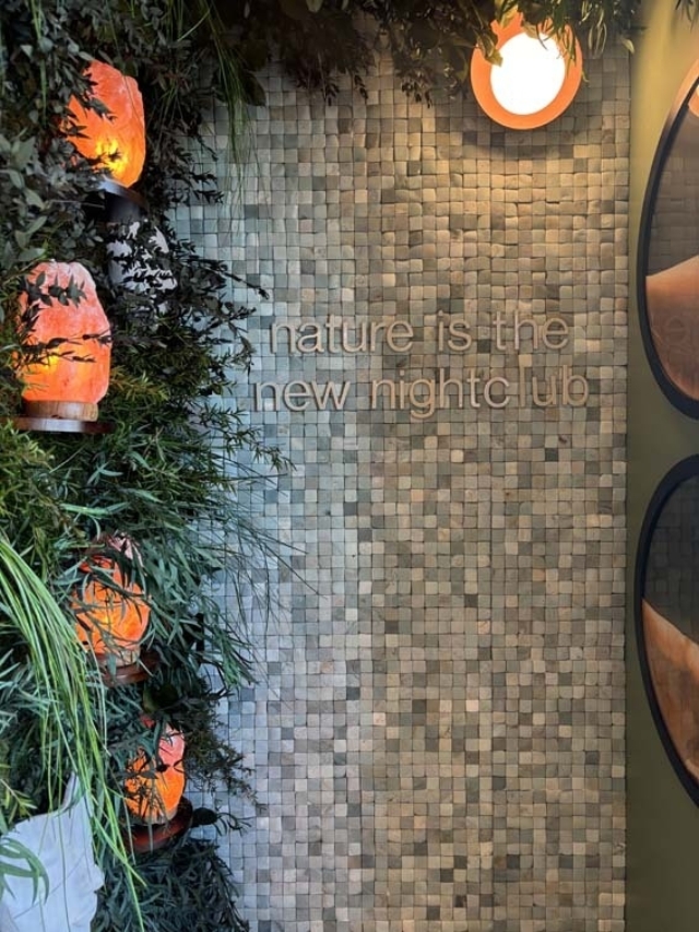 Mosaic tile wall with metal letters reading 'nature is the new nightclub', tall green plants and glowing amber salt lamps along left side.