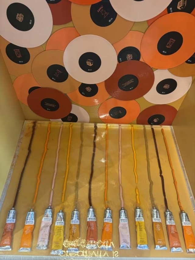 Metal paint tubes drip vertical orange, brown, yellow and pink toward a ceiling of overlapping orange and white vinyl records.