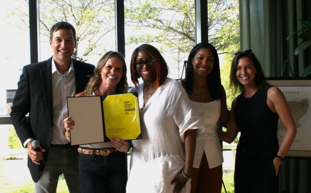 Five smiling adults stand by a large window at an awards event, one woman holding a framed yellow IIDA Winner certificate while others pose.