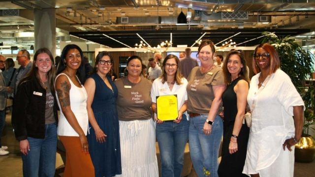 Nine diverse women stand shoulder-to-shoulder in a modern office, smiling while one woman holds a yellow award plaque, celebratory mood.