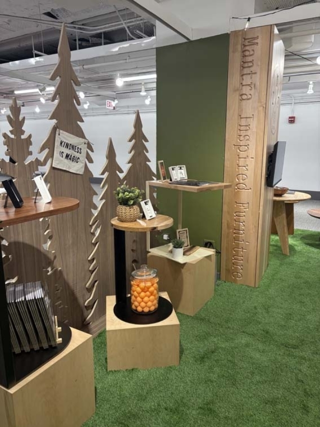 Showroom with wooden tree cutouts, pedestals and shelves on green turf, a jar of orange balls and a 'Kindness is Magic' sign.