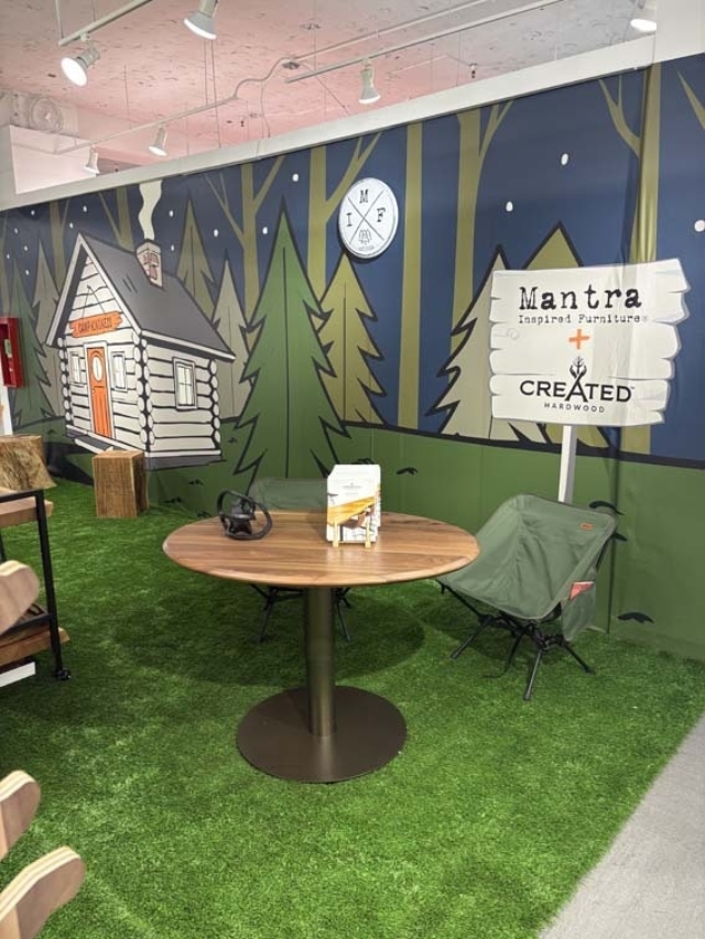 Indoor showroom styled as a forest with cabin mural, faux grass floor, round table with brochure and headphones and a green camping chair.