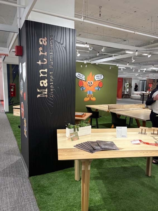 Bright modern showroom with wood tables on green turf, a black pillar reading Mantra, pamphlets on a table and an orange leaf mascot mural.