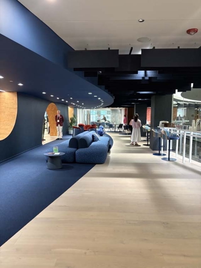 Modern open lounge with curved navy ceiling, modular blue sofas on dark carpet, light wood floors, bar stools to the right and people.
