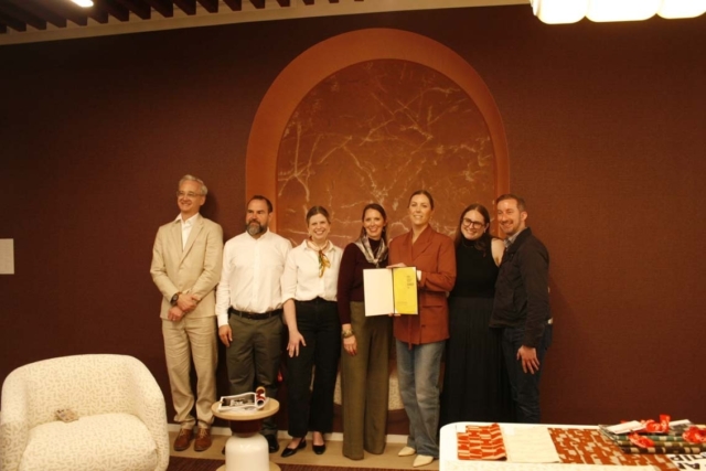 Seven people stand in a warm, brown room beneath an arched marble niche, smiling as a woman near center holds a yellow certificate.