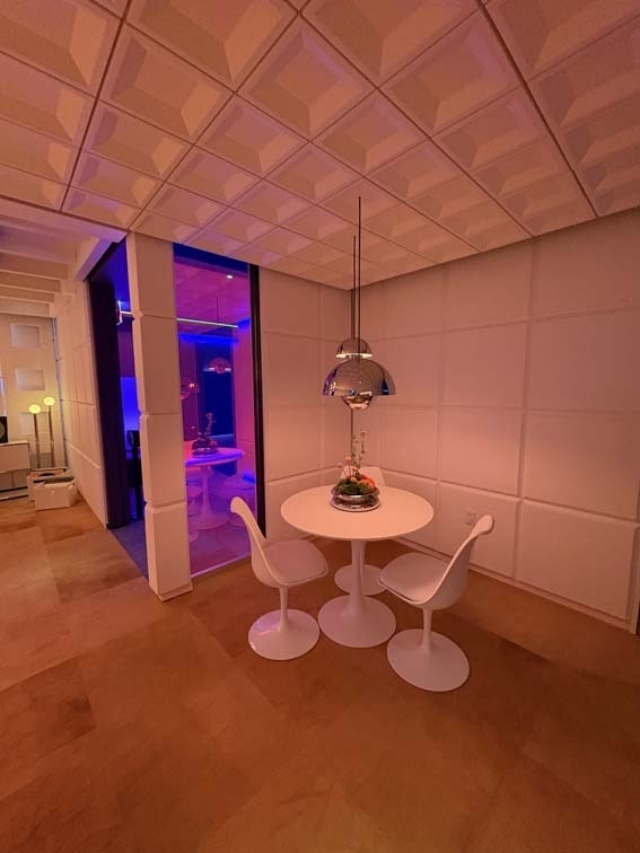 Retro-futuristic dining nook with round white table and two tulip chairs under a chrome dome pendant, bathed in pink-orange light.