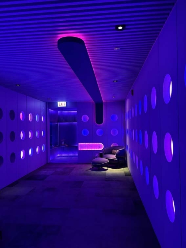 A futuristic neon-lit corridor with purple and pink circular wall cutouts, recessed ceiling fixture, lounge chair and EXIT sign at far end.