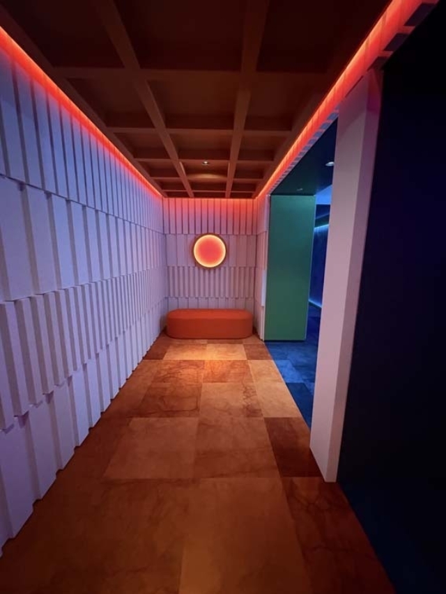Narrow modern hallway with textured panels, amber tiled floor, red LED strip lighting, orange circular wall light above a rounded bench.