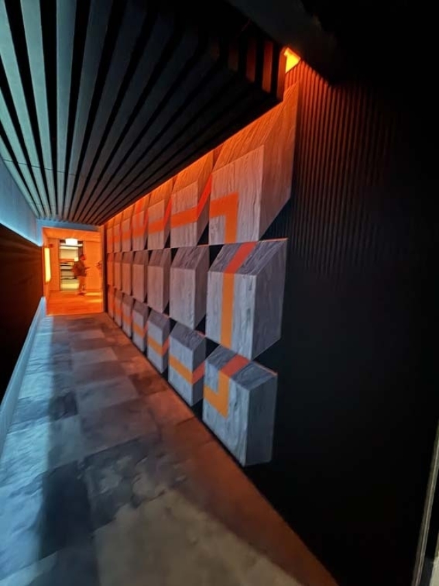 Narrow modern corridor with gray cubic wall panels edged in orange, slatted ceiling, blue ambient lighting and a warm doorway glow ahead.