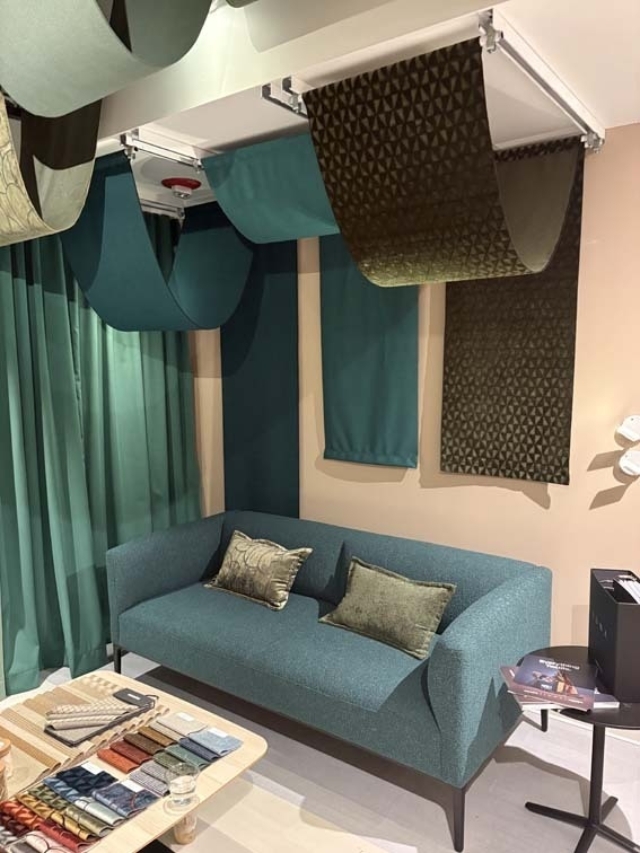 Teal sofa with two green patterned cushions beneath hanging teal and olive fabric swatches, curtains, and a sample table nearby.