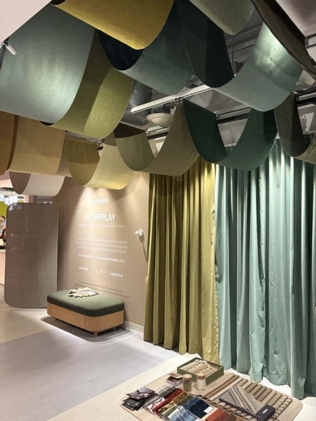 Showroom corner with hanging green and yellow fabric waves, long curtains, a cushioned bench and swatches laid out on a table.