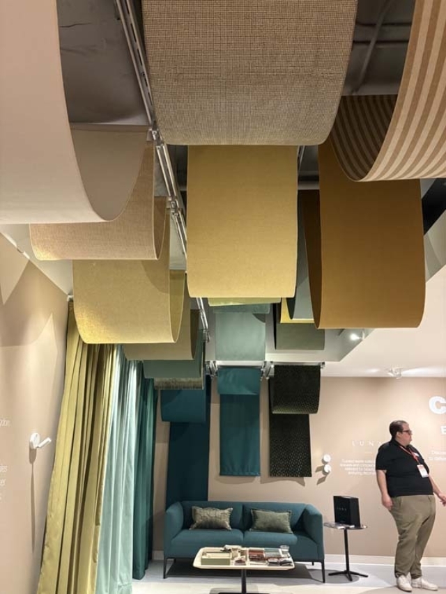 Showroom with large suspended fabric rolls in mustard, teal and beige above a teal sofa, green curtains, coffee table with books and a man.
