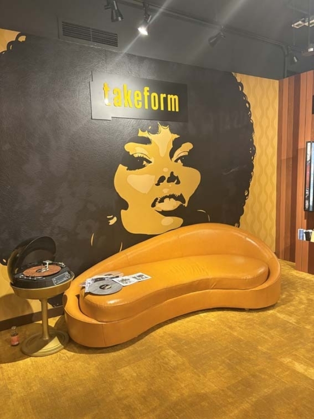 Curvy orange sofa beside a spinning record player and magazine below a black-and-gold mural of an Afro woman's face with a 'takeform' sign.