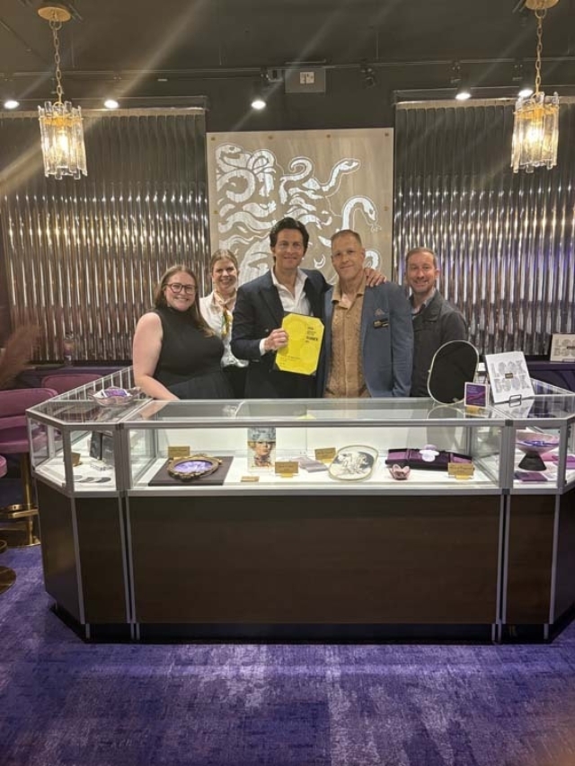 Five smiling staff stand behind a glass case of jewelry, purple carpet and metallic octopus wall art; center man holds a yellow bag.
